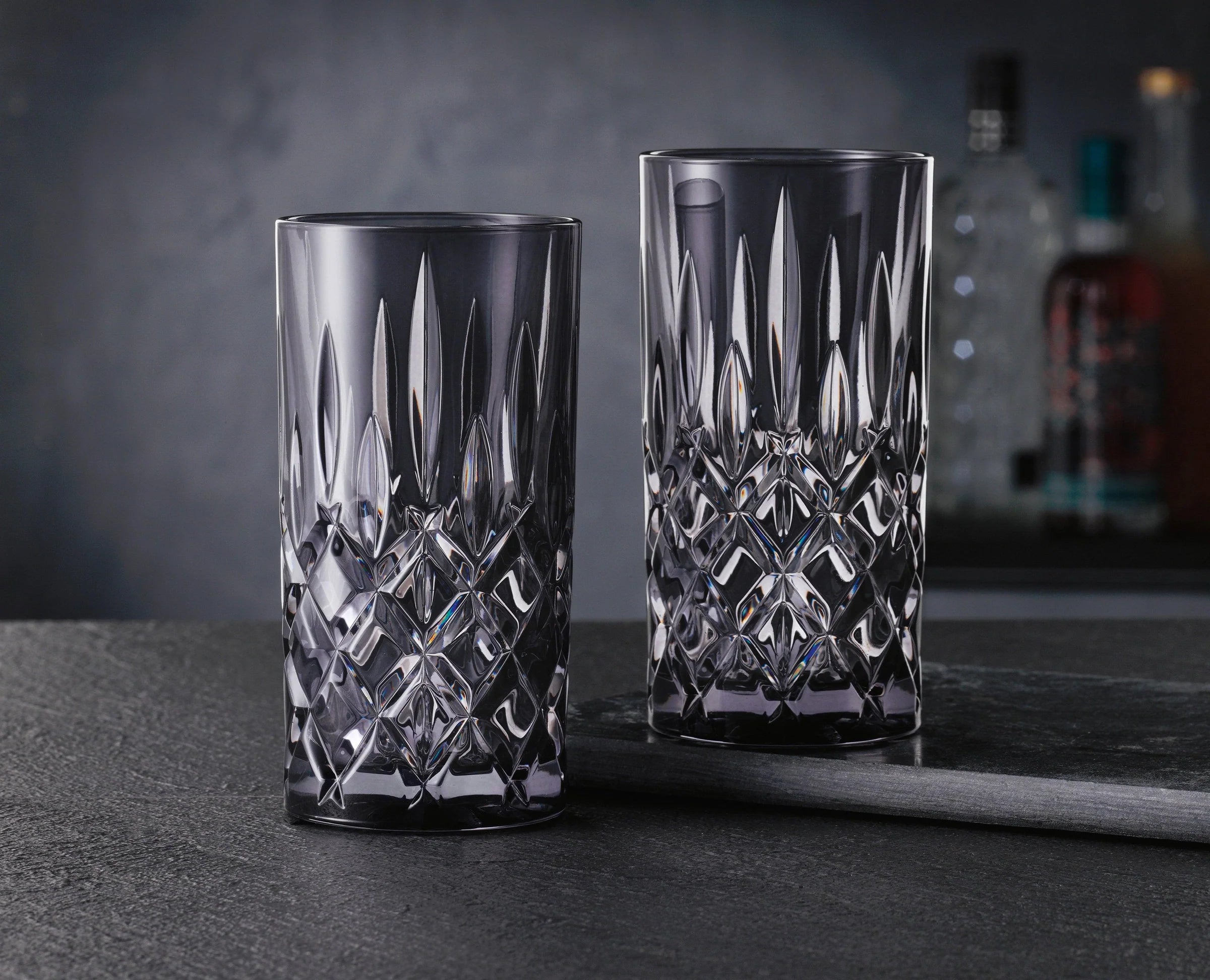 Two crystal glasses with intricate designs on a dark surface, blurred bottles in the background.