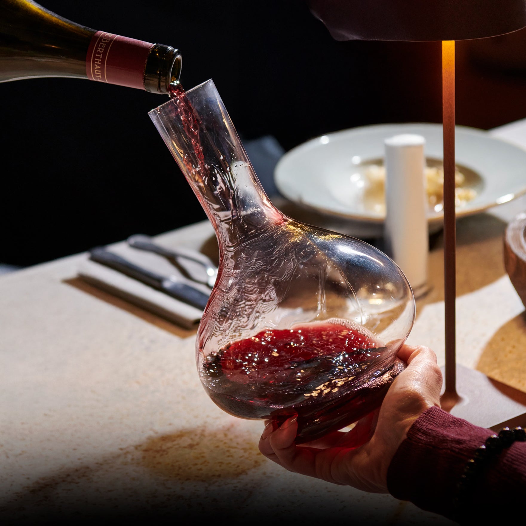 Red wine being poured into a glass decanter on a dining table.