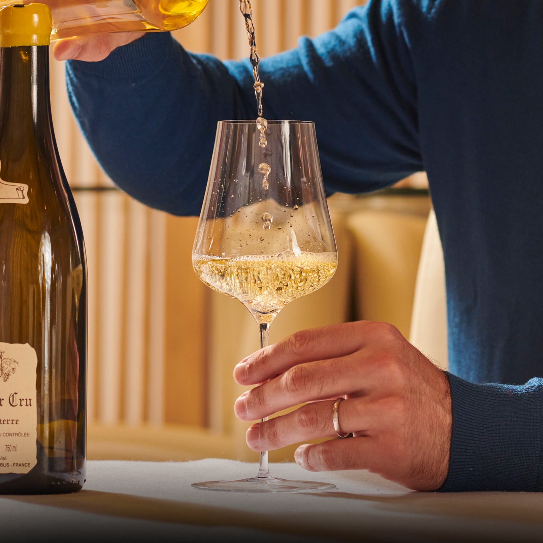 Person pouring white wine into a glass with a bottle of wine in the background.
